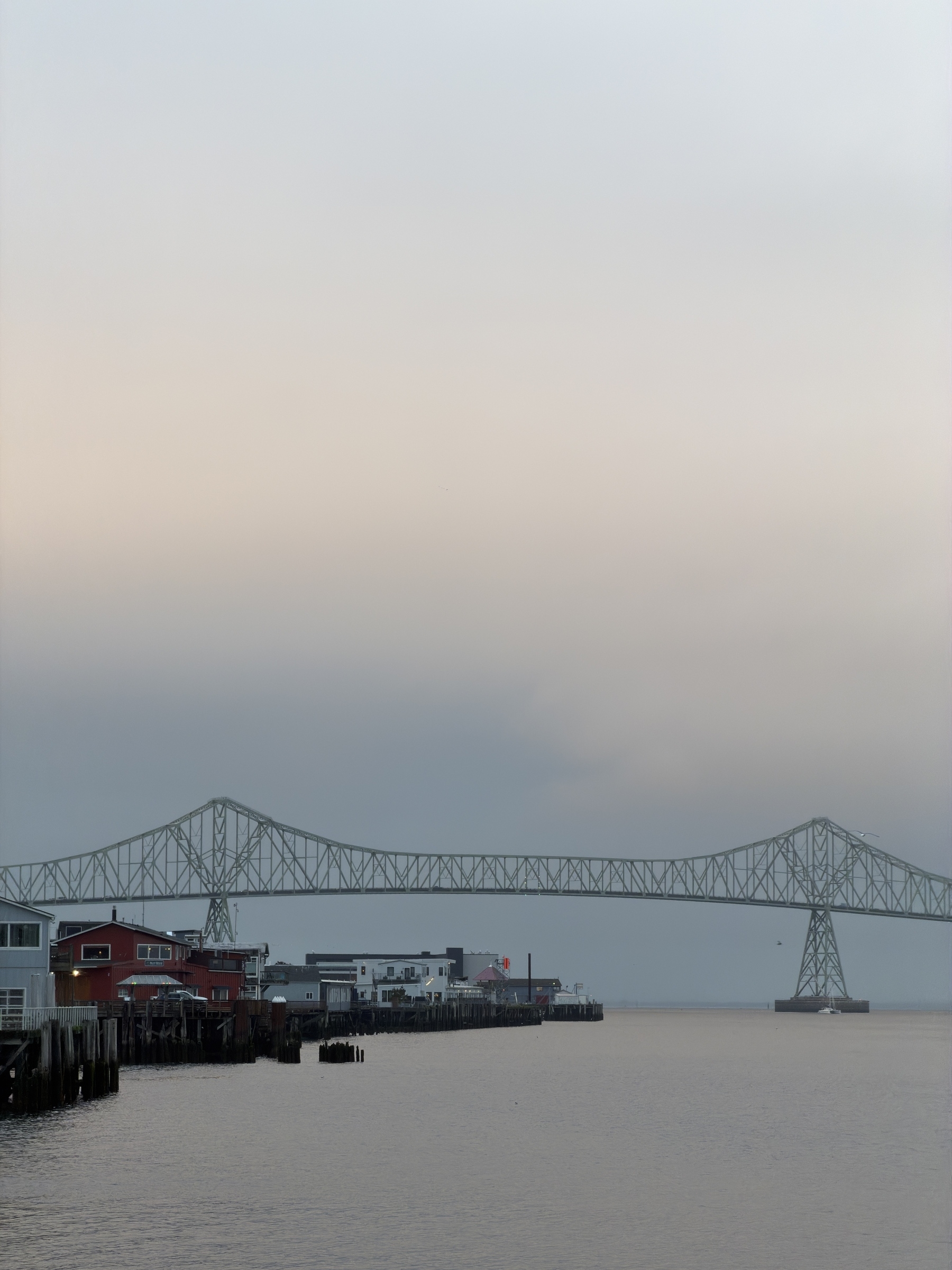 A large bridge spans across a calm body of water near a dock with buildings under a cloudy sky.