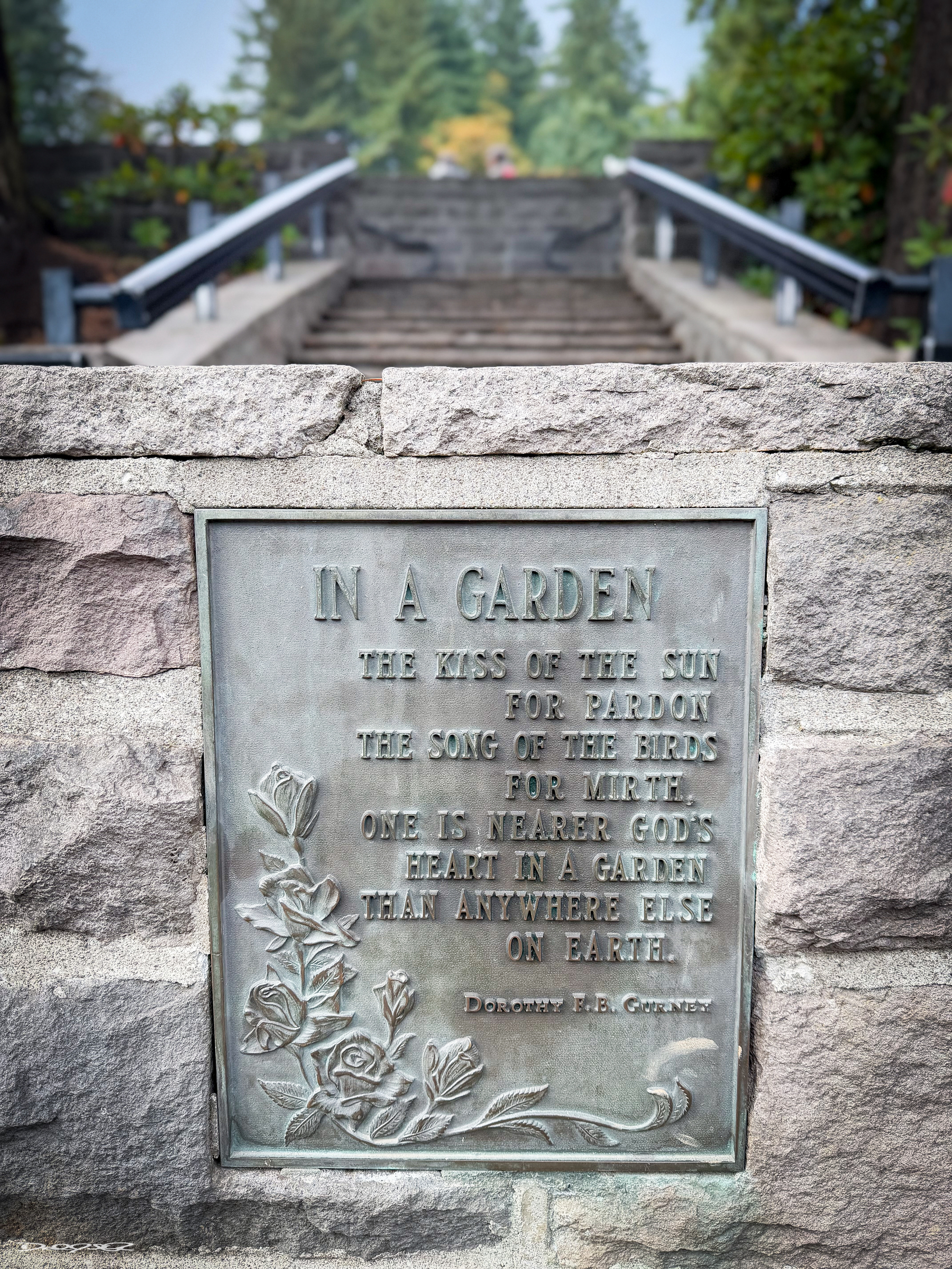 A stone wall features a plaque with a poem about the spiritual beauty of gardens, set in a lush outdoor setting with a staircase and trees in the background.