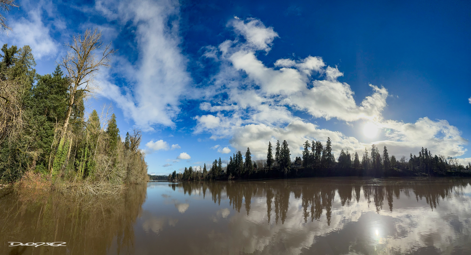 A calm river reflects a vibrant blue sky with scattered clouds, bordered by lush trees.