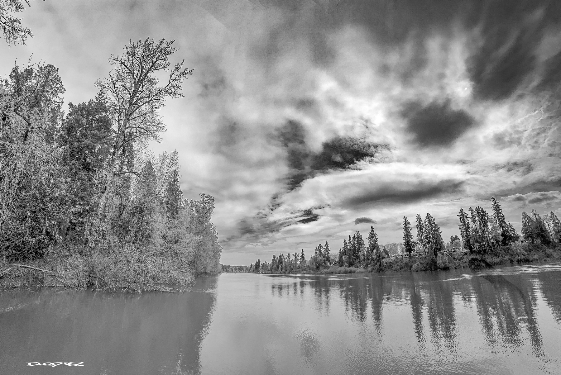 A serene Willamette River scene showcases calm water reflecting the surrounding trees and a dramatic, cloud-filled sky.