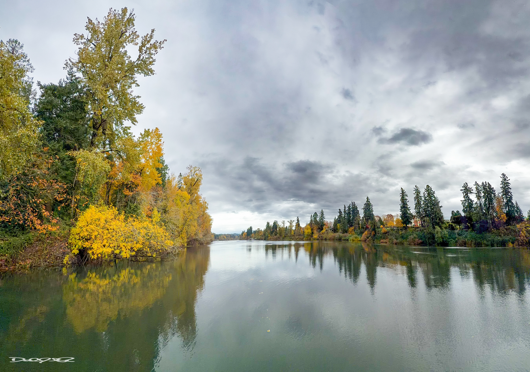 A serene river is flanked by autumn-colored trees under a cloudy sky.