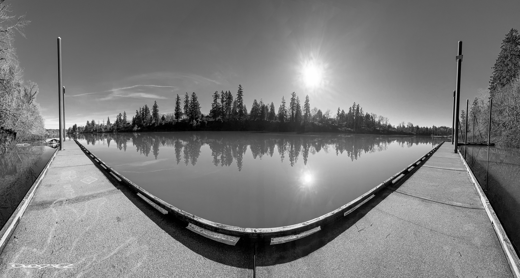 A wide-angle, black and white view of a serene river with a dock extending into the water, surrounded by trees and under a bright sun.