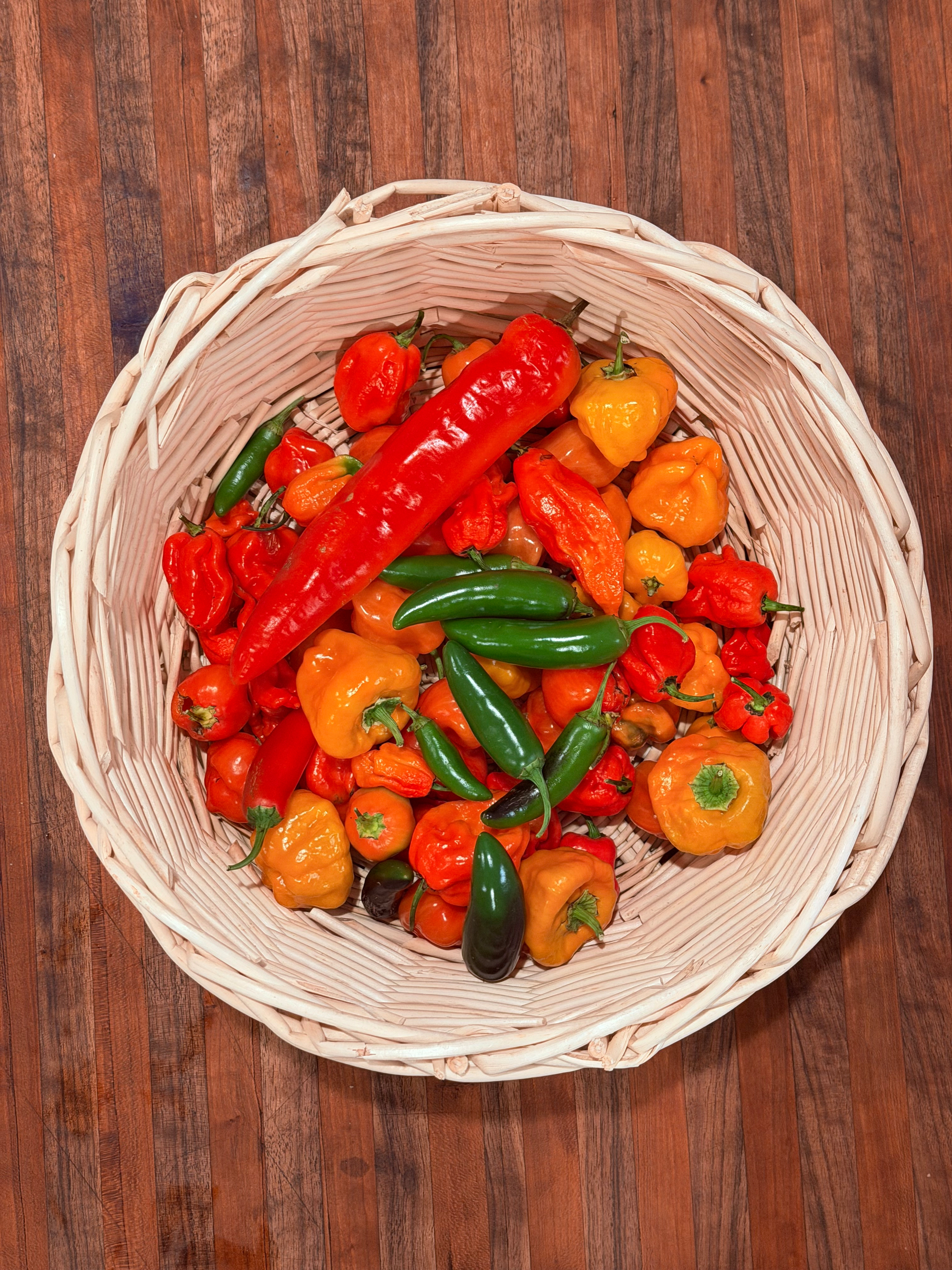 A basket filled with a variety of colorful peppers is placed on a wooden surface.