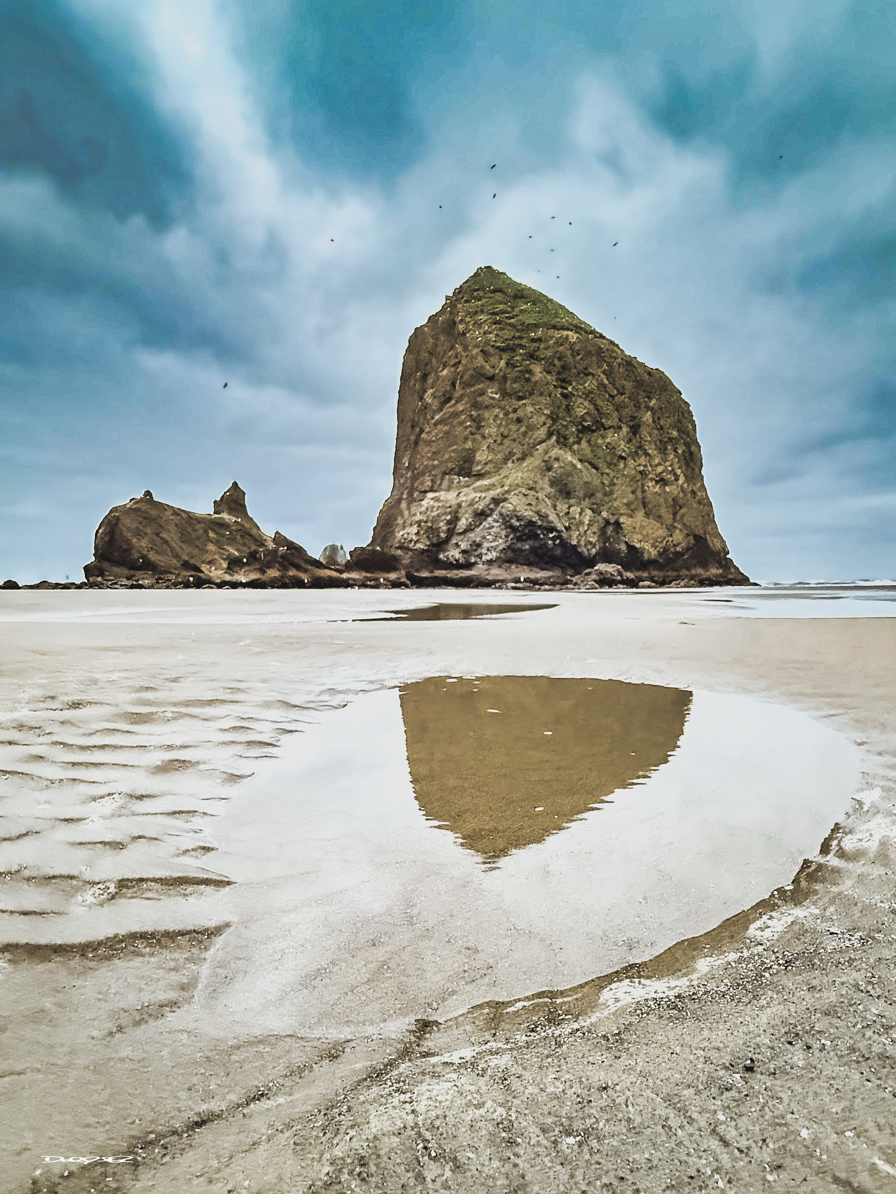 A large rock formation called Haystack Rock rises from the sandy beach with a reflective tide pool in the foreground under a cloudy sky at dawn.