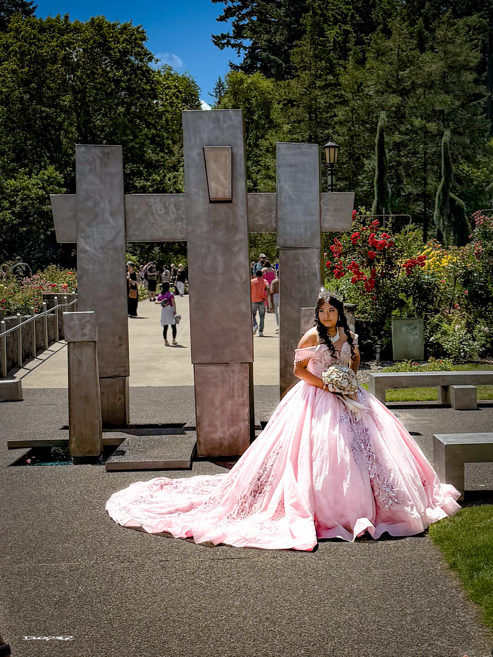 A young woman in a pink gown holds a bouquet while posing in a garden with large metal sculptures and trees in the background.