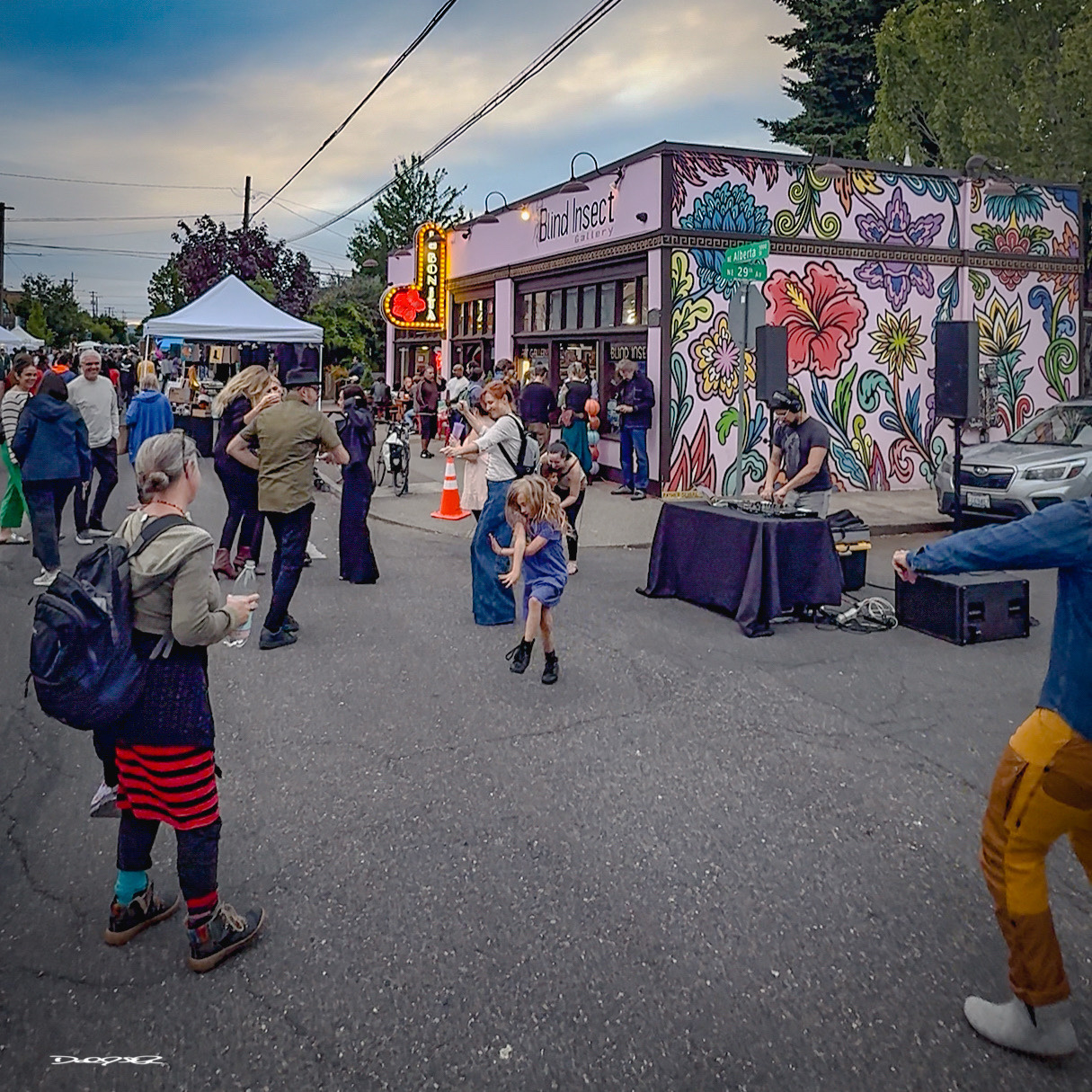 People are gathered at an outdoor event with street art, music & DJ, as children play and others socialize.