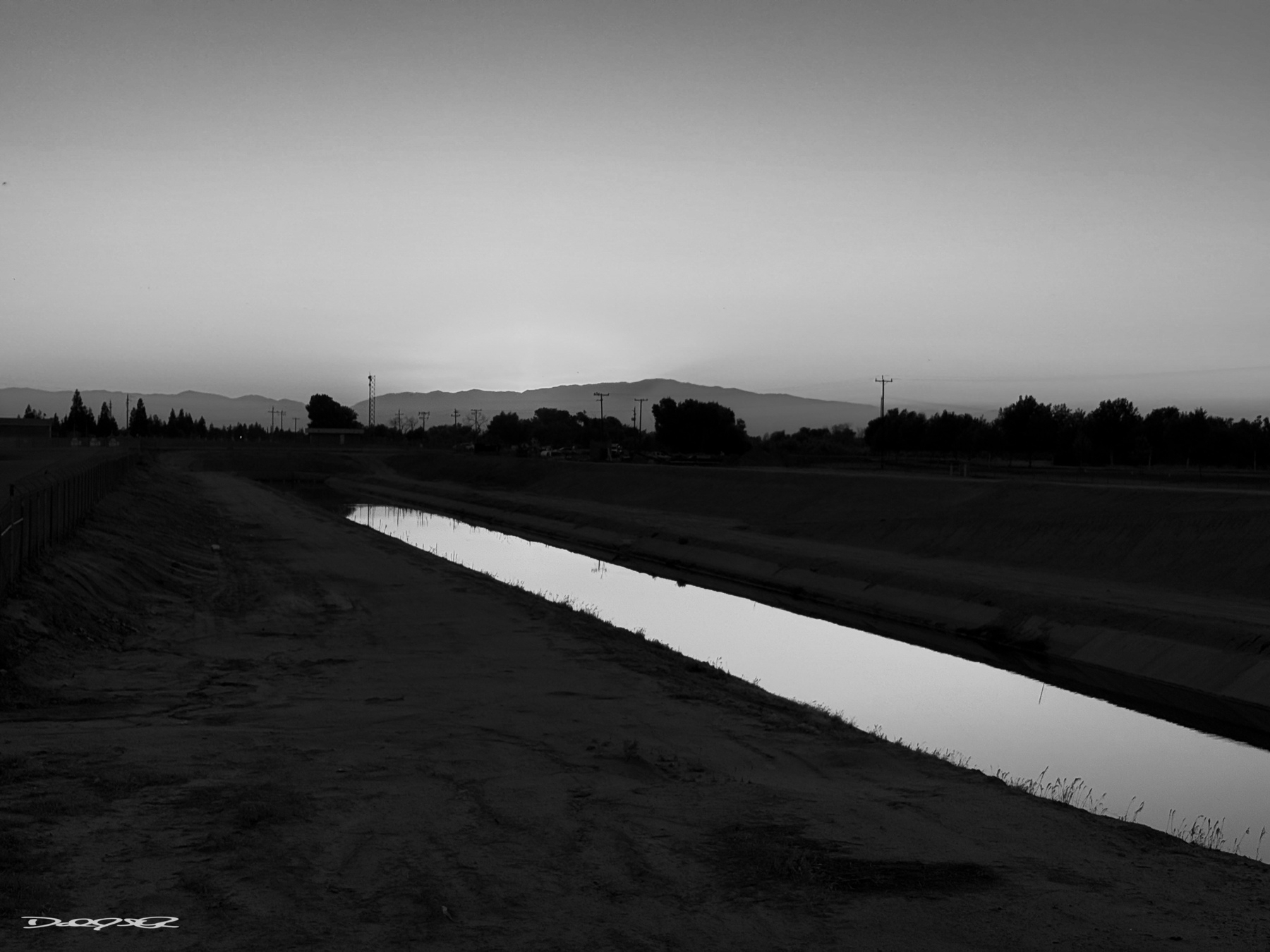A serene black and white landscape features a reflective canal running through a flat, expansive terrain with silhouetted trees and mountains in the distance under a softly fading sky.