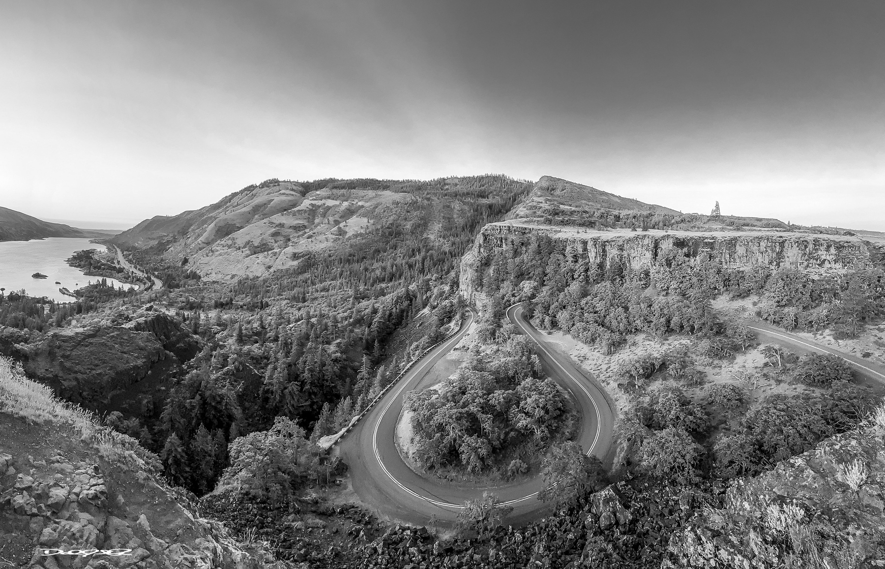 A winding road snakes through a lush mountainous landscape under a clear sky, with a river visible on the left.