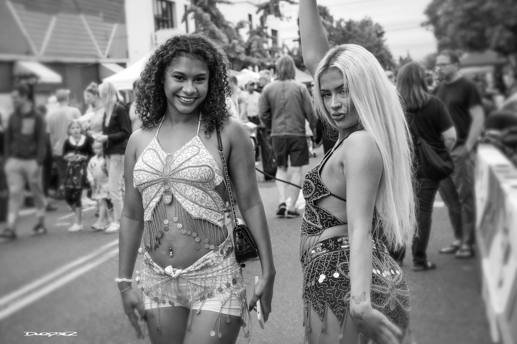 Two women in festive costumes pose for the camera during an outdoor event, surrounded by people.