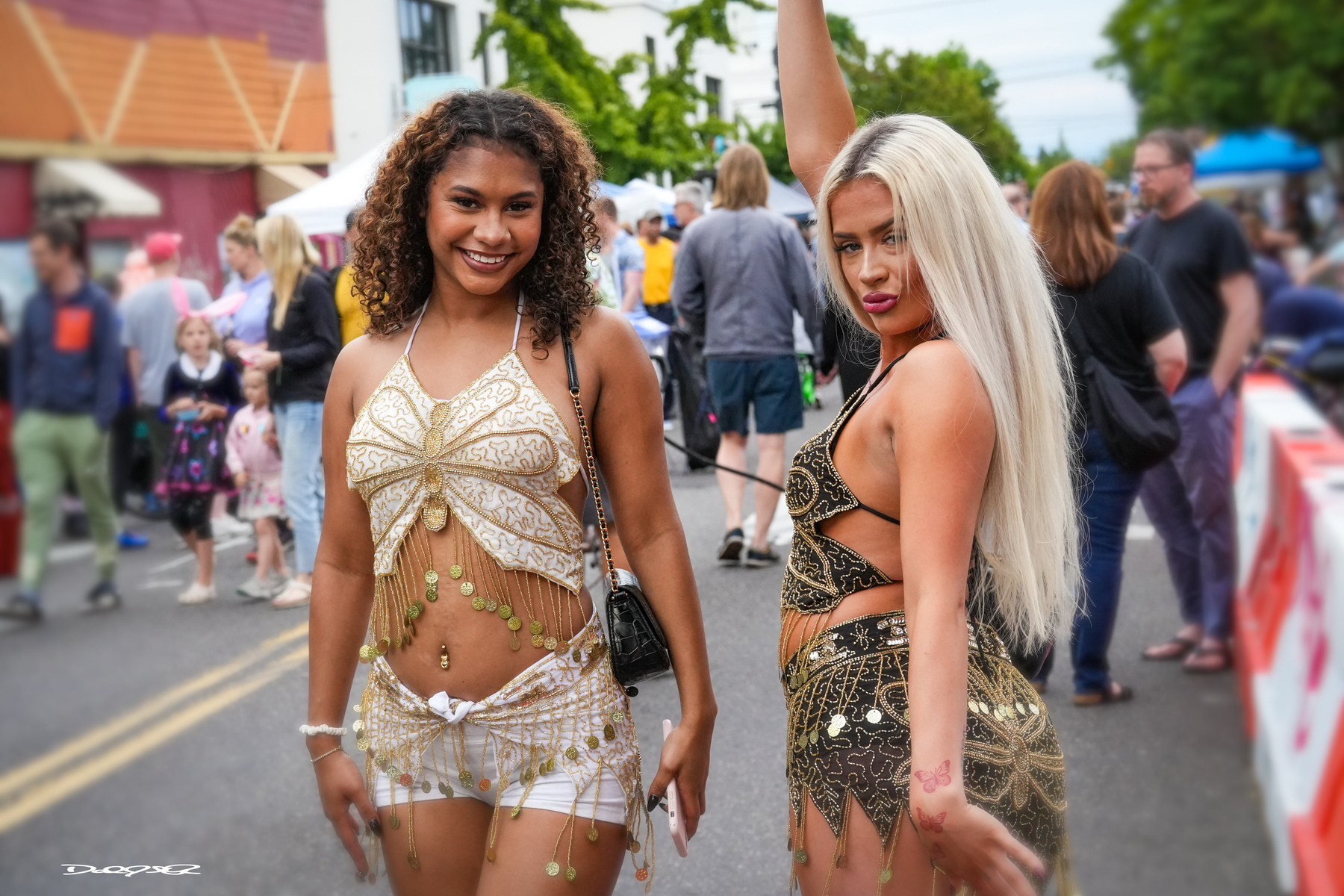 Two women in distinctive, embellished outfits pose confidently on a busy outdoor street with onlookers in the background.