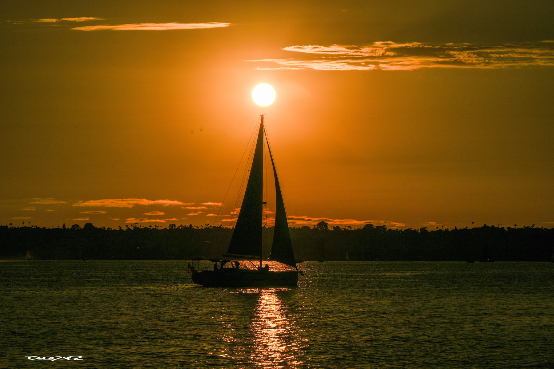 A sailboat glides across the water under a stunning orange sunset with the sun perfectly positioned above the horizon.