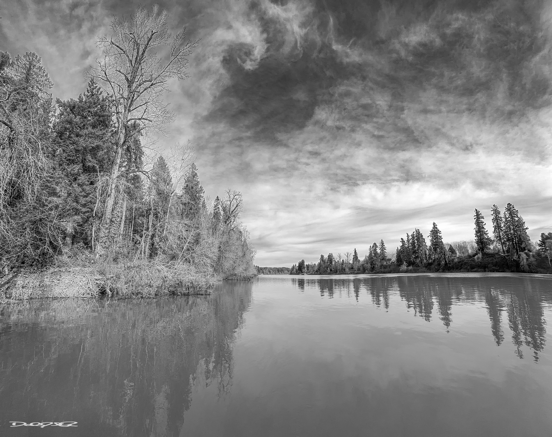 A serene black and white landscape features a calm Willame River flanked by trees under a dramatic, cloudy sky.