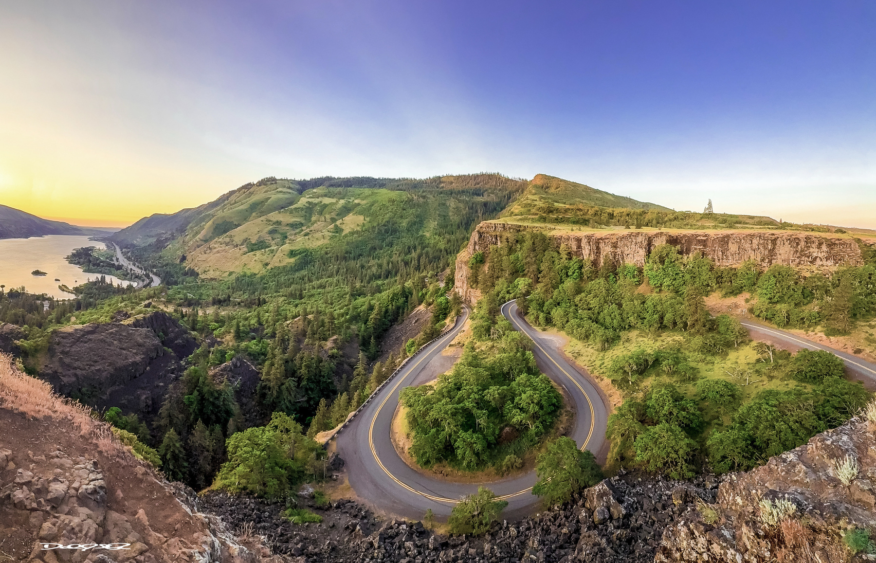 A winding road curves through a lush, hilly landscape with a river visible in the background under a clear sky.