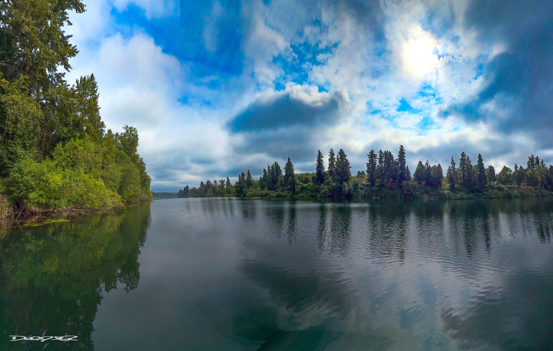 A serene river landscape is bordered by lush green trees under a partly cloudy blue sky.