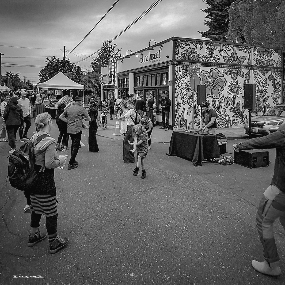 A lively street scene features people gathering near a colorful mural-covered building with a musician performing at a small outdoor event.