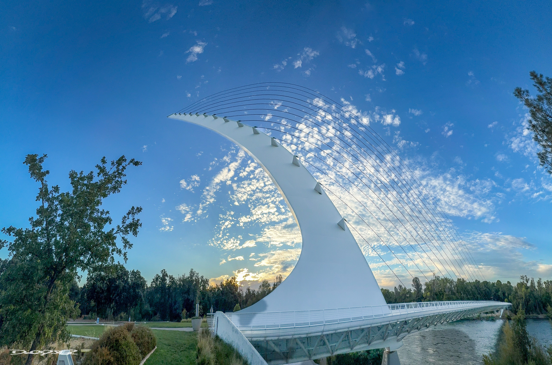 A striking white, modern sundial bridge stretches over a river against a backdrop of a blue sky with scattered clouds and surrounding trees.