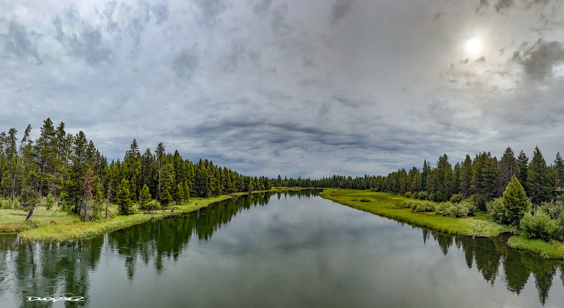 A serene Deschutes River flows through a lush, forested landscape under a cloudy sky with the sun peeking through.