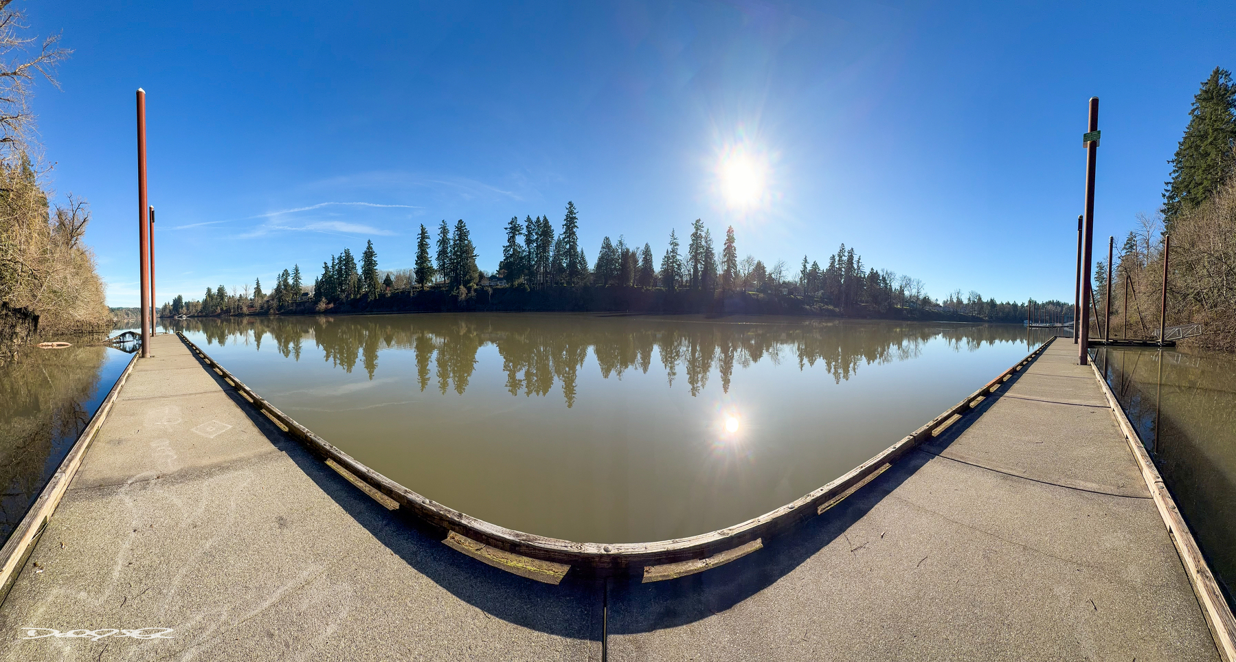 A panoramic view of a calm river with a dock, surrounded by trees under a clear blue sky and the sun shining brightly.