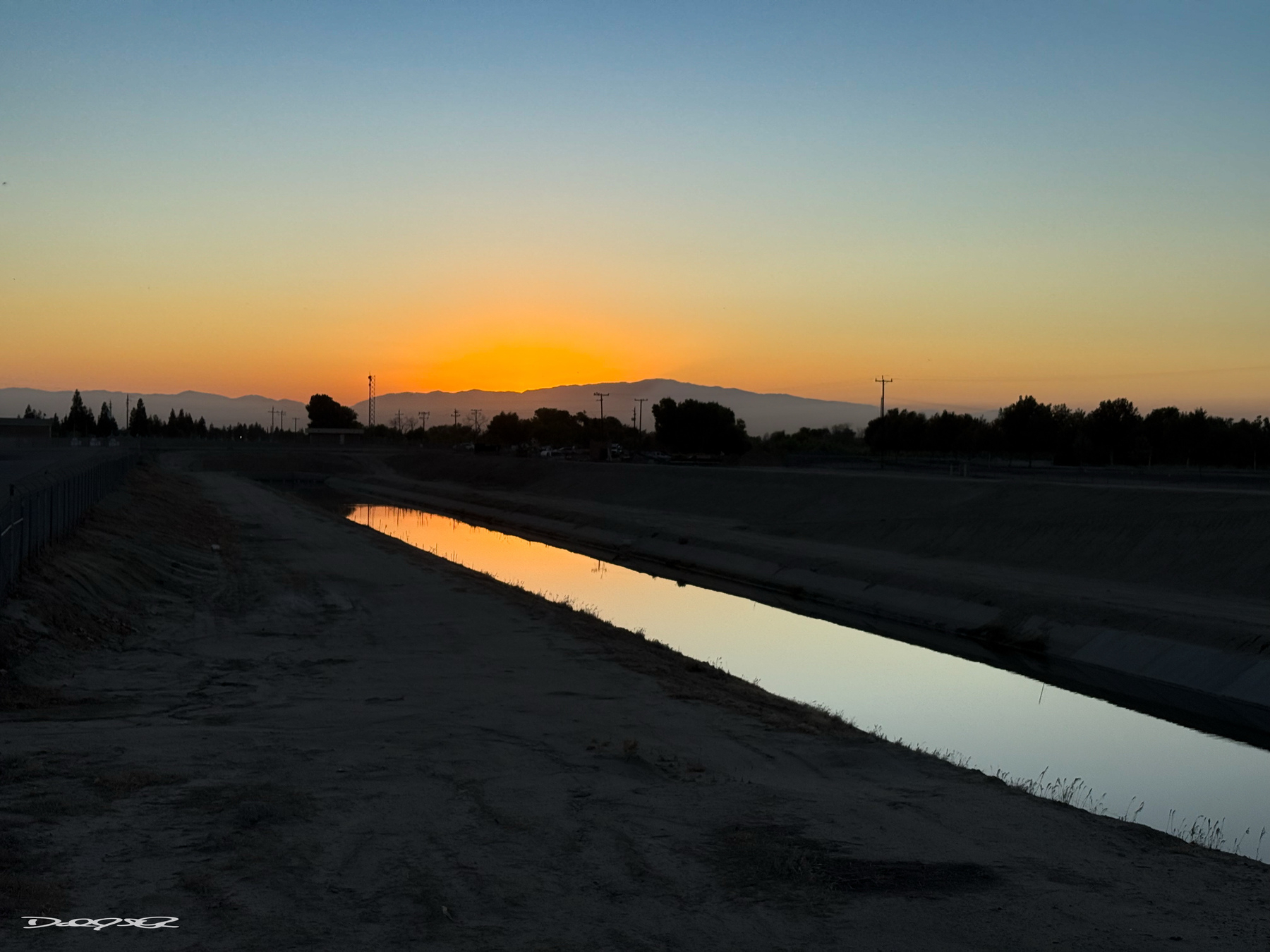 A serene sunset casts a warm glow over a narrow canal with silhouetted trees and distant mountains.