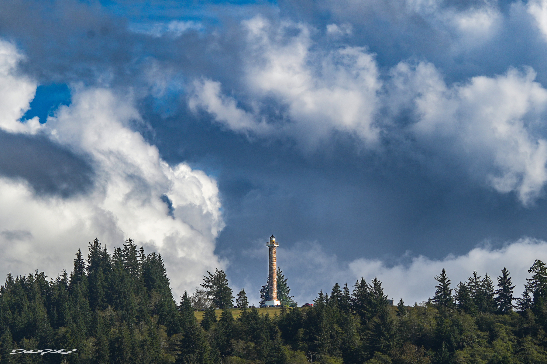 The Astoria Column, surrounded by a forest, is set against a backdrop of dramatic, cloudy skies.