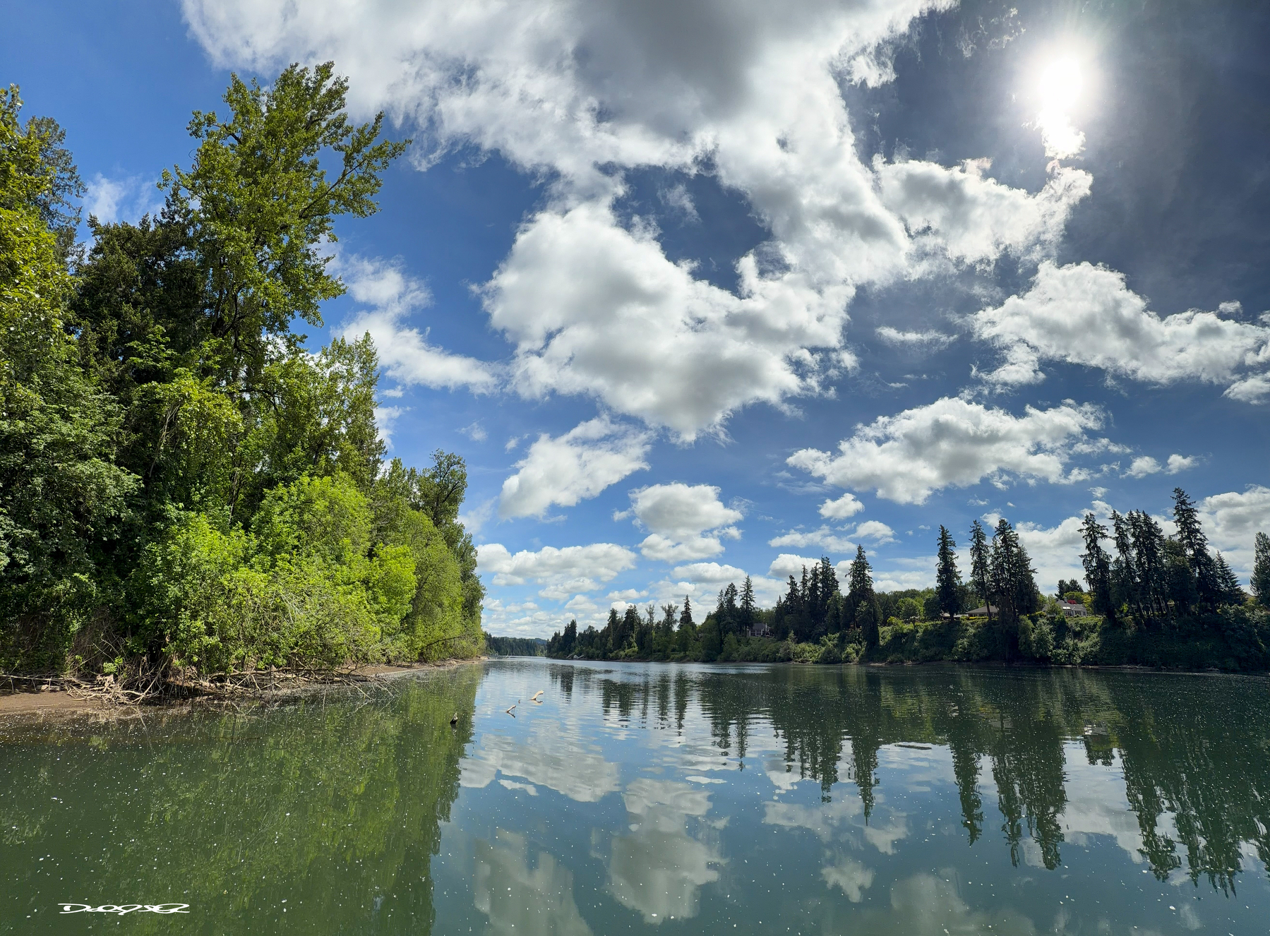 A serene river scene features calm reflective waters, lush green trees, and a partly cloudy sky with sunlight.