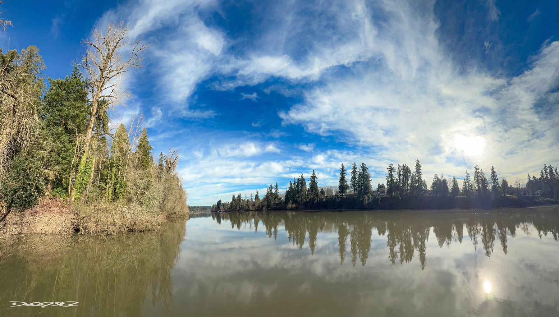 A serene river reflects a clear sky and distant trees on a bright, sunny day.