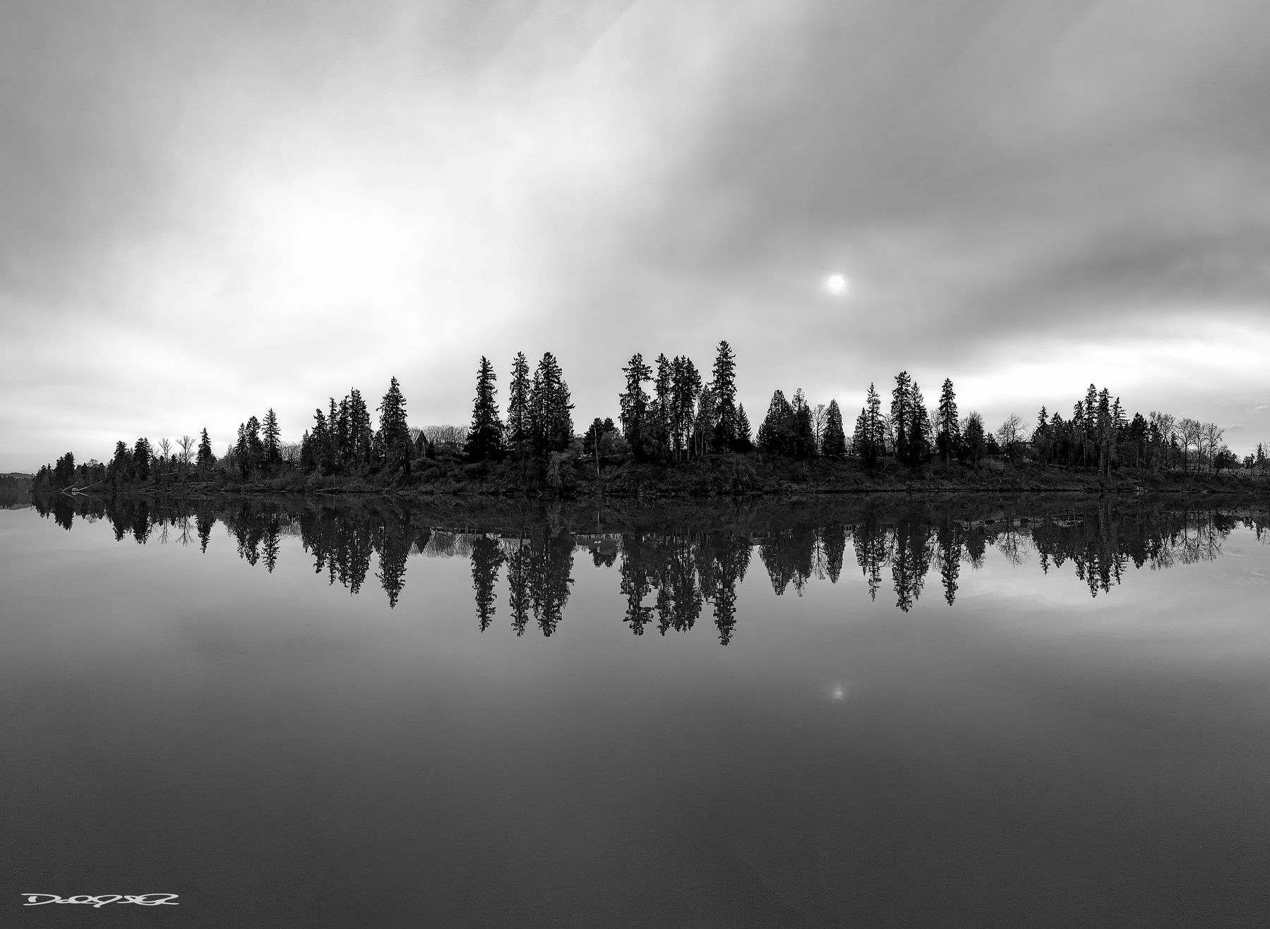 A serene, black and white landscape features trees reflecting perfectly on the still surface of a river under a cloudy sky.