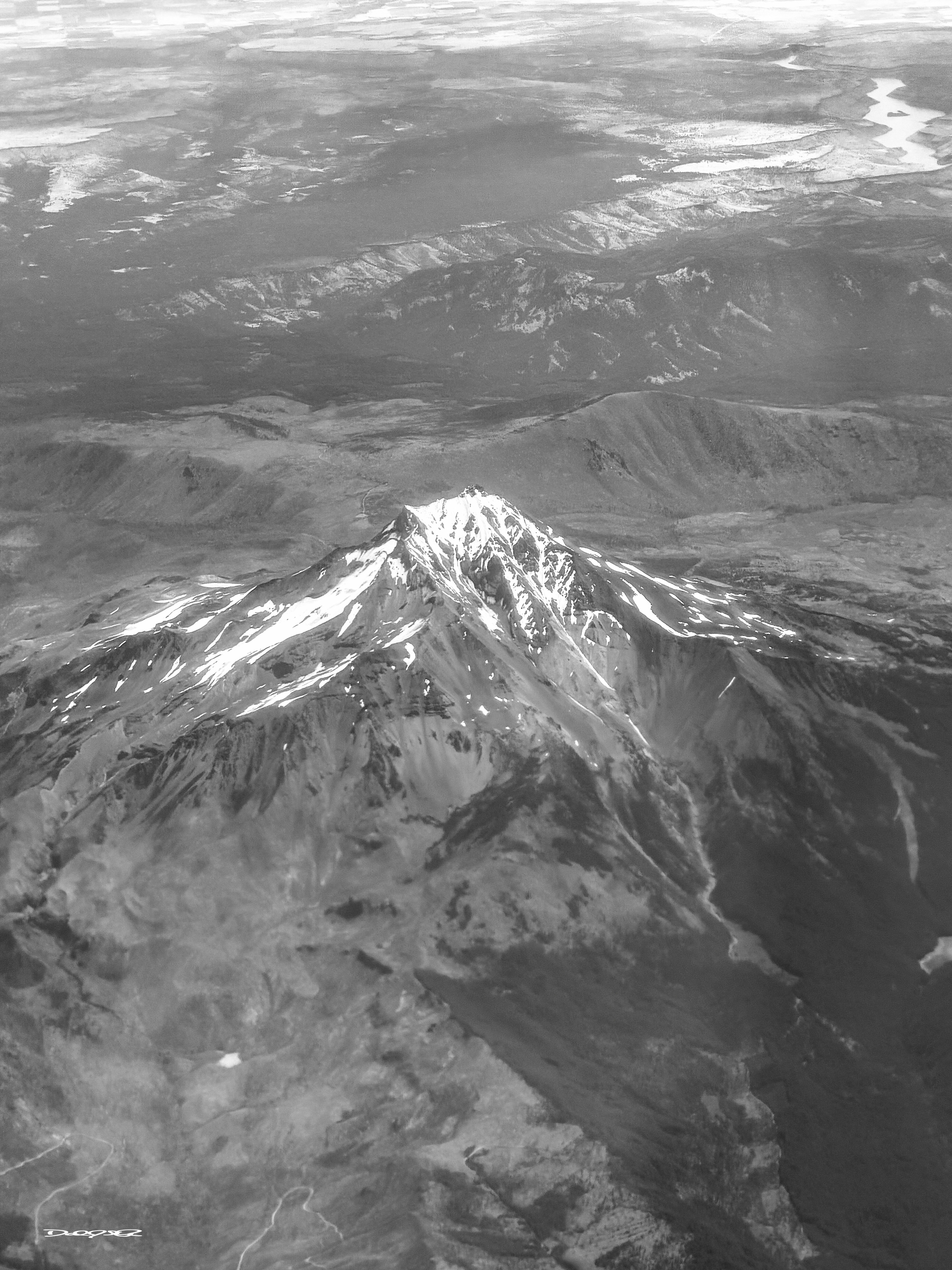 A snow-capped mountain peak rises prominently against a backdrop of rugged terrain and distant bodies of water.