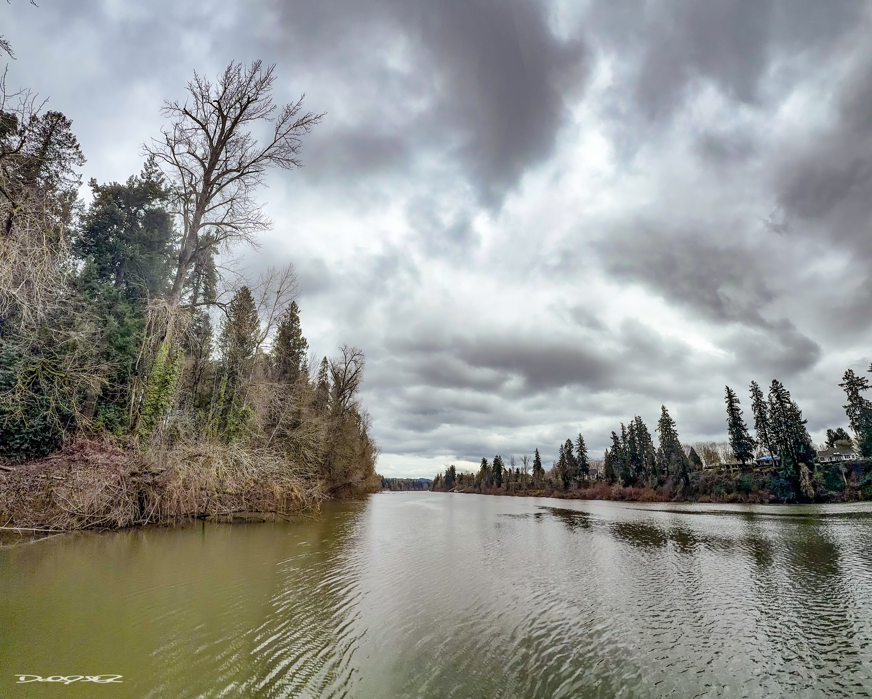 A serene river flows under a cloudy sky, bordered by trees on both banks.