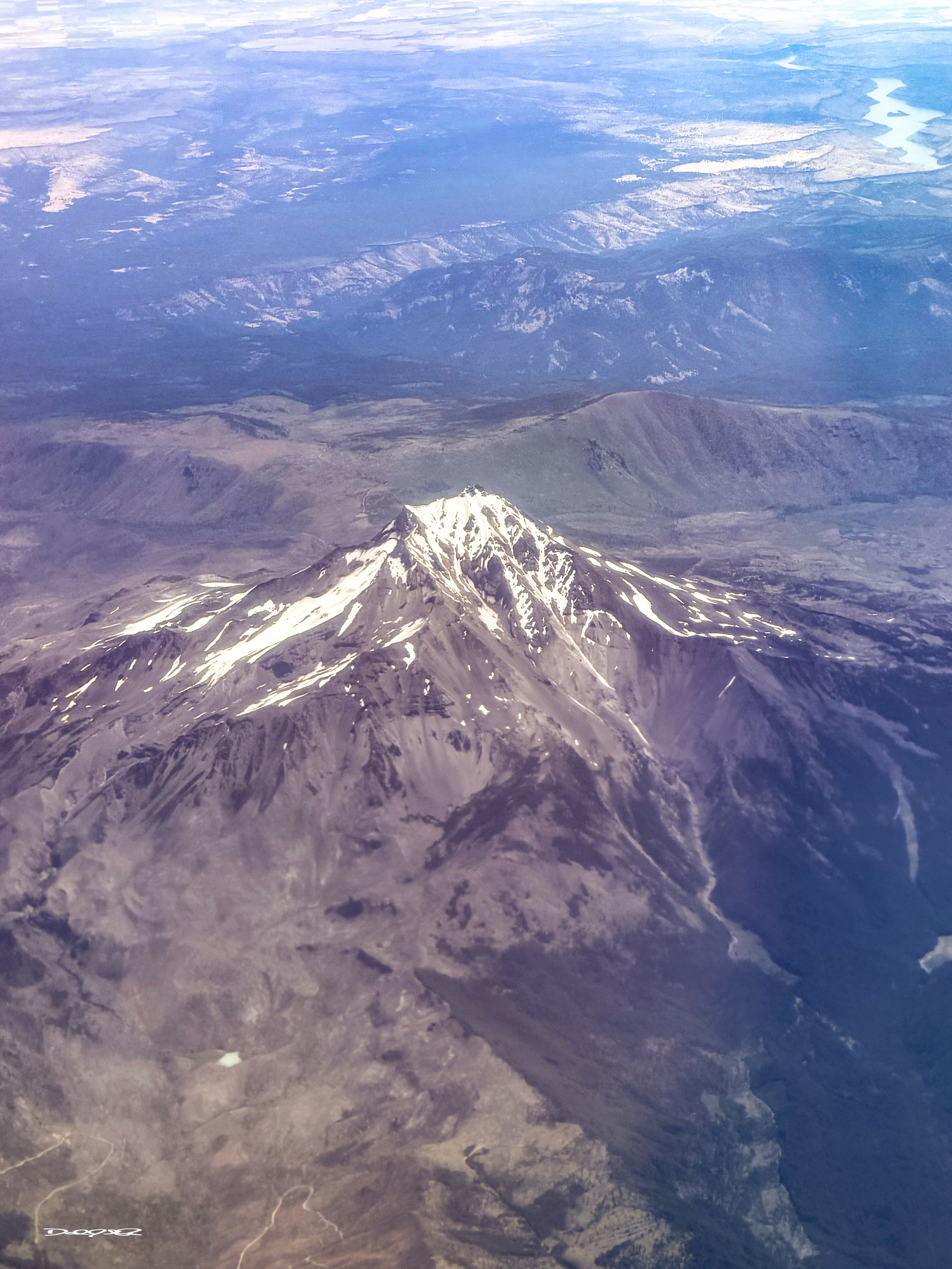 A snow-capped mountain peak rises prominently above a rugged, expansive landscape with distant hills and a winding river visible in the background.