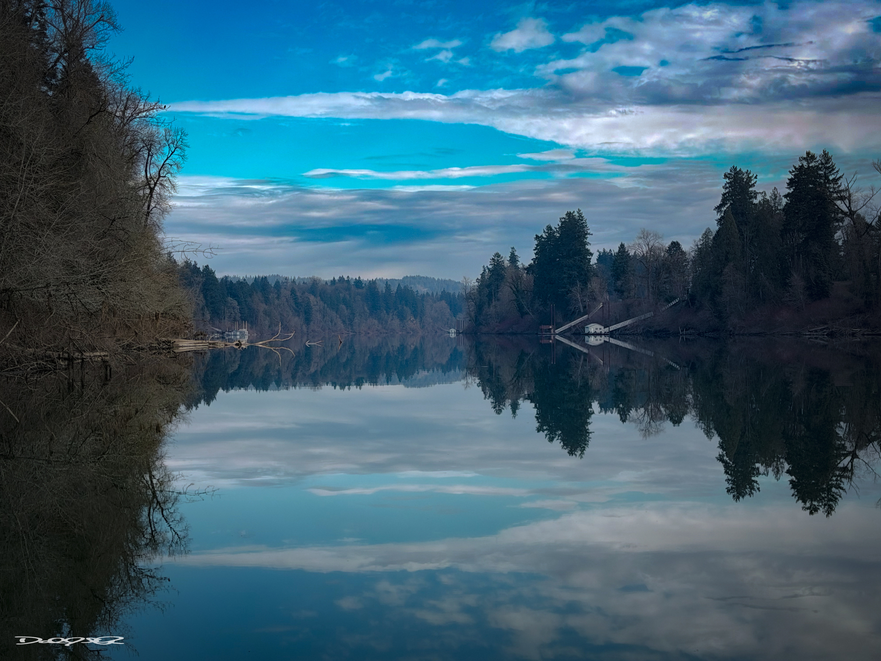 A tranquil river is flanked by trees on both sides, with reflections of the sky and clouds on the water's surface.