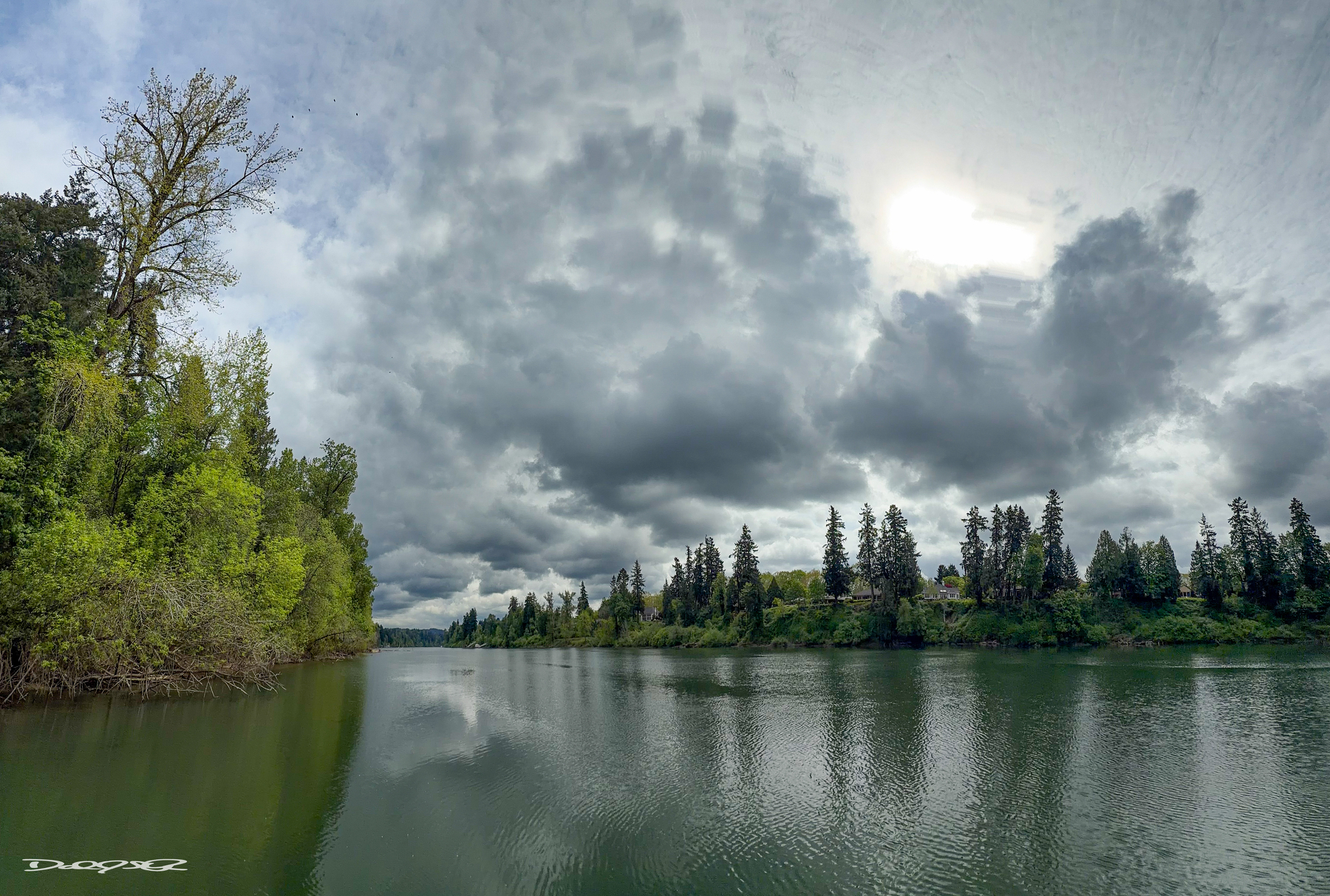 A calm Willamette River is surrounded by lush green trees under a cloudy sky with the sun peeking through.