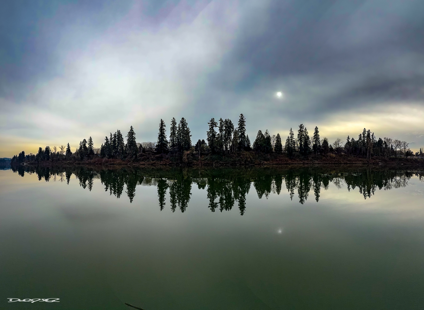 A tranquil river reflects a dense line of evergreen trees under a cloudy sky with a faint sun.